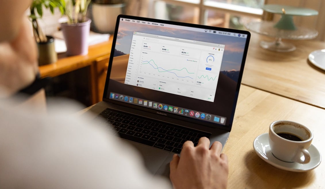 Photograph from the perspective of a person sitting at a desk looking at their laptop with graphs and charts on the screen. A cup of coffee is beside it.