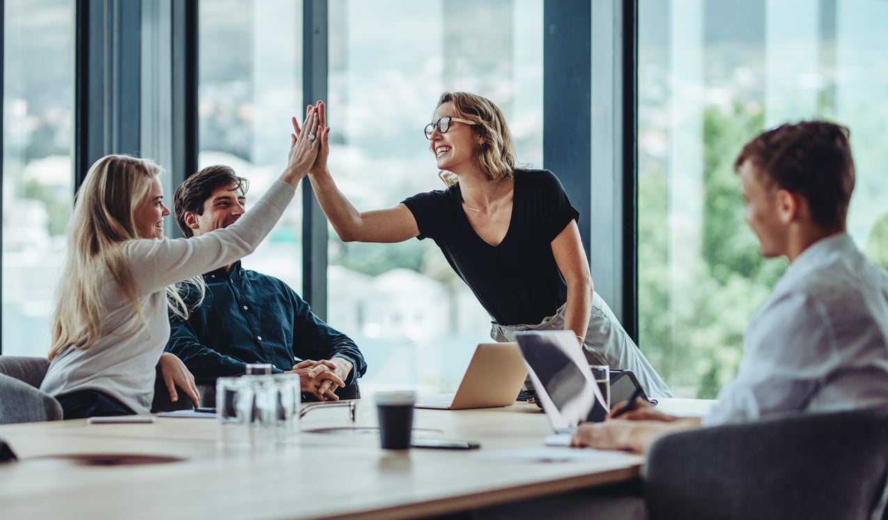 photo of a team of four people at an office conference table. all are smiling and two women are high-fiving one another.