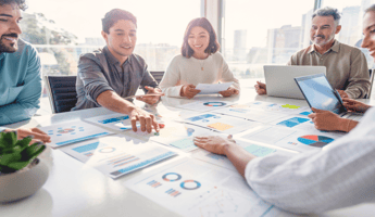 Business people sitting at a table looking at printed out graphs and charts. They are all smiling and appear to be collaborating with one another.
