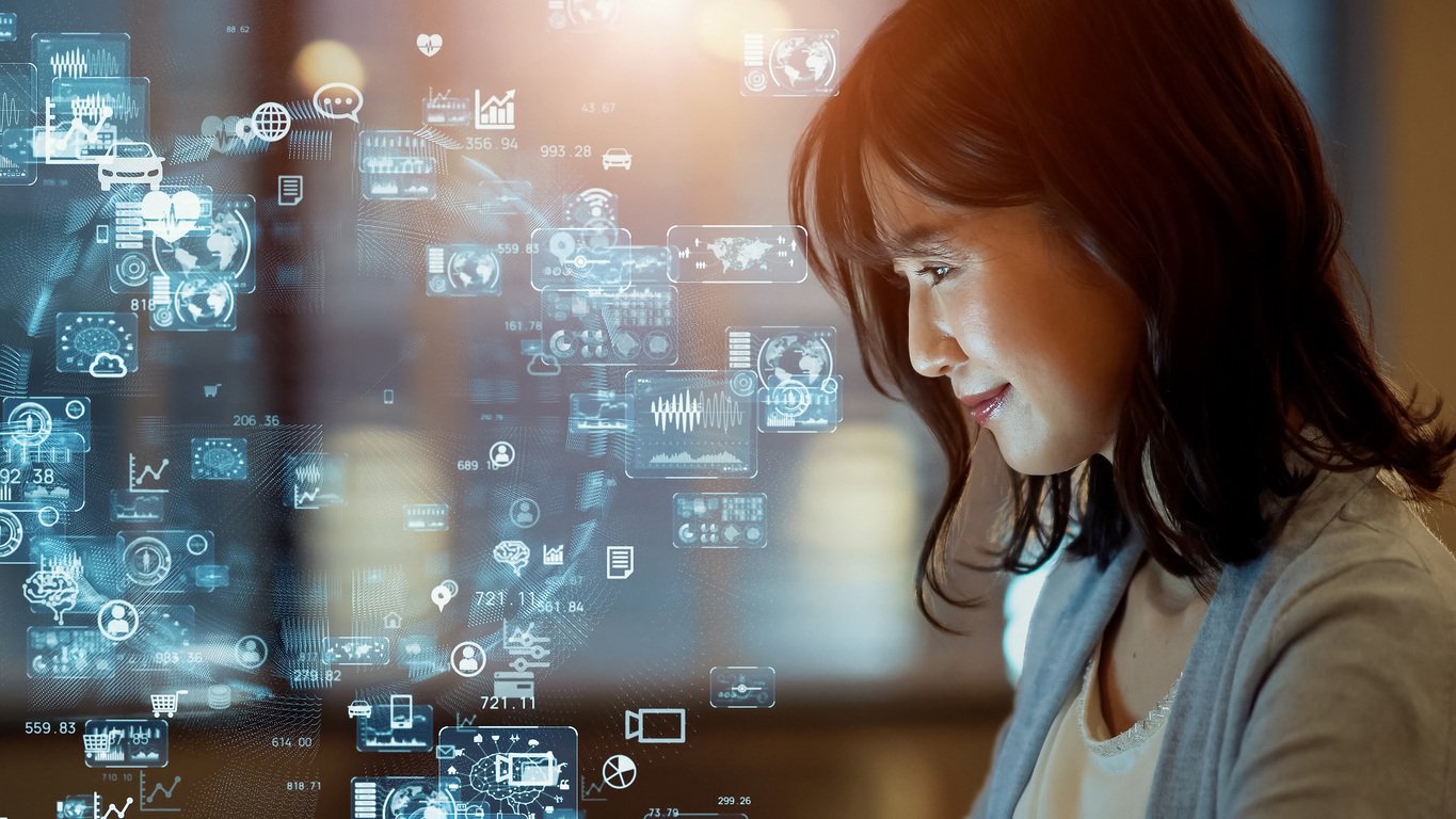 A woman with a small smile looking at her computer. Overlayed on the image are tiny transparent icons of graphs and charts and lines indicating how they are all connected.